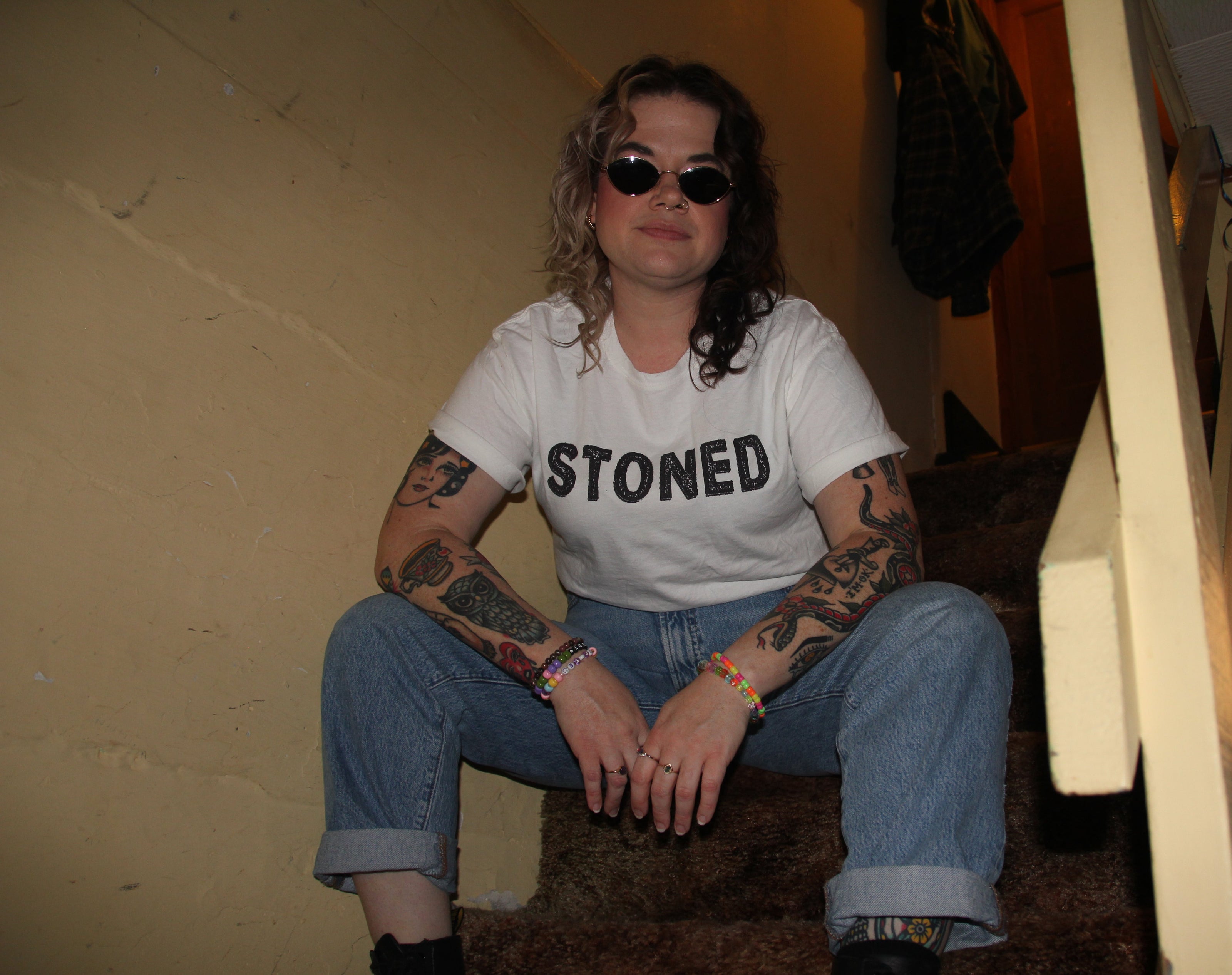 Person wearing a 'STONED' t-shirt sitting on a step indoors.